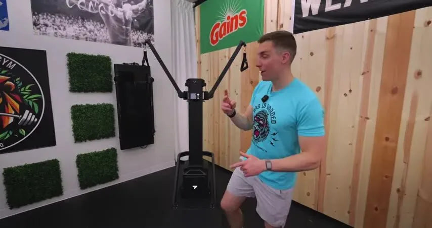 Man demonstrating FitTransformer Titan cable machine in room with wood wall, green hedge tiles, and 'Gains' banner.